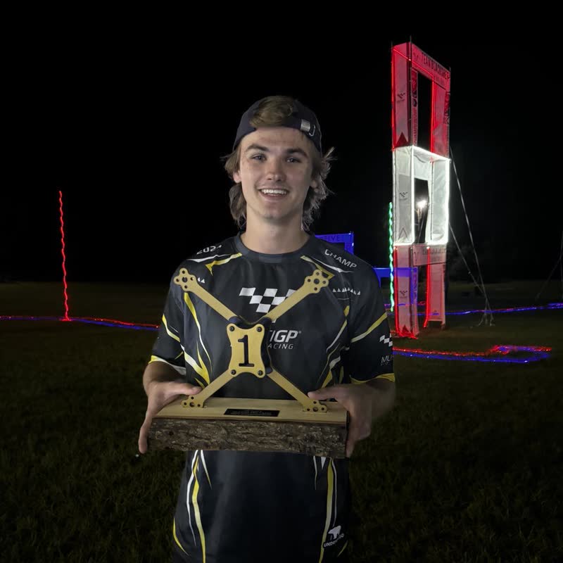 Paweł Laszczak nocą z pucharem MultiGP Championship — złotą jedynką na drewnianej podstawie.